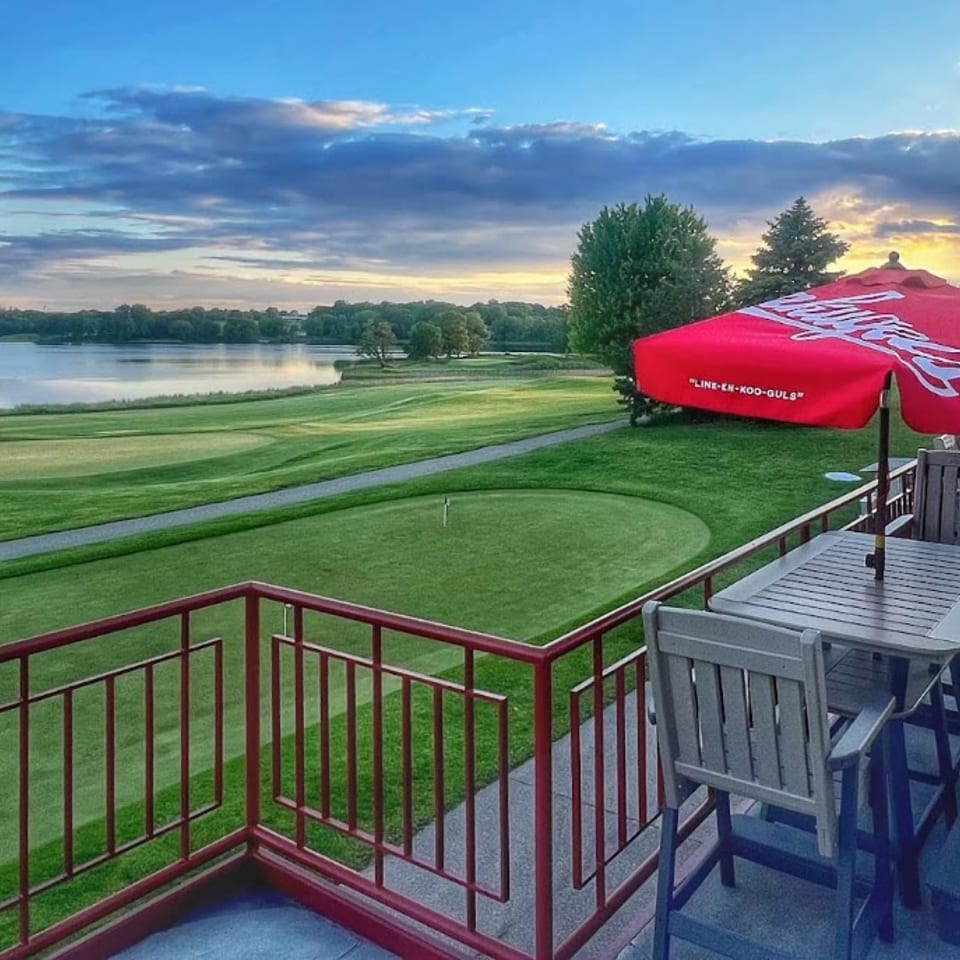 Patio dining area with red umbrellas overlooking a golf course and lake at sunset.