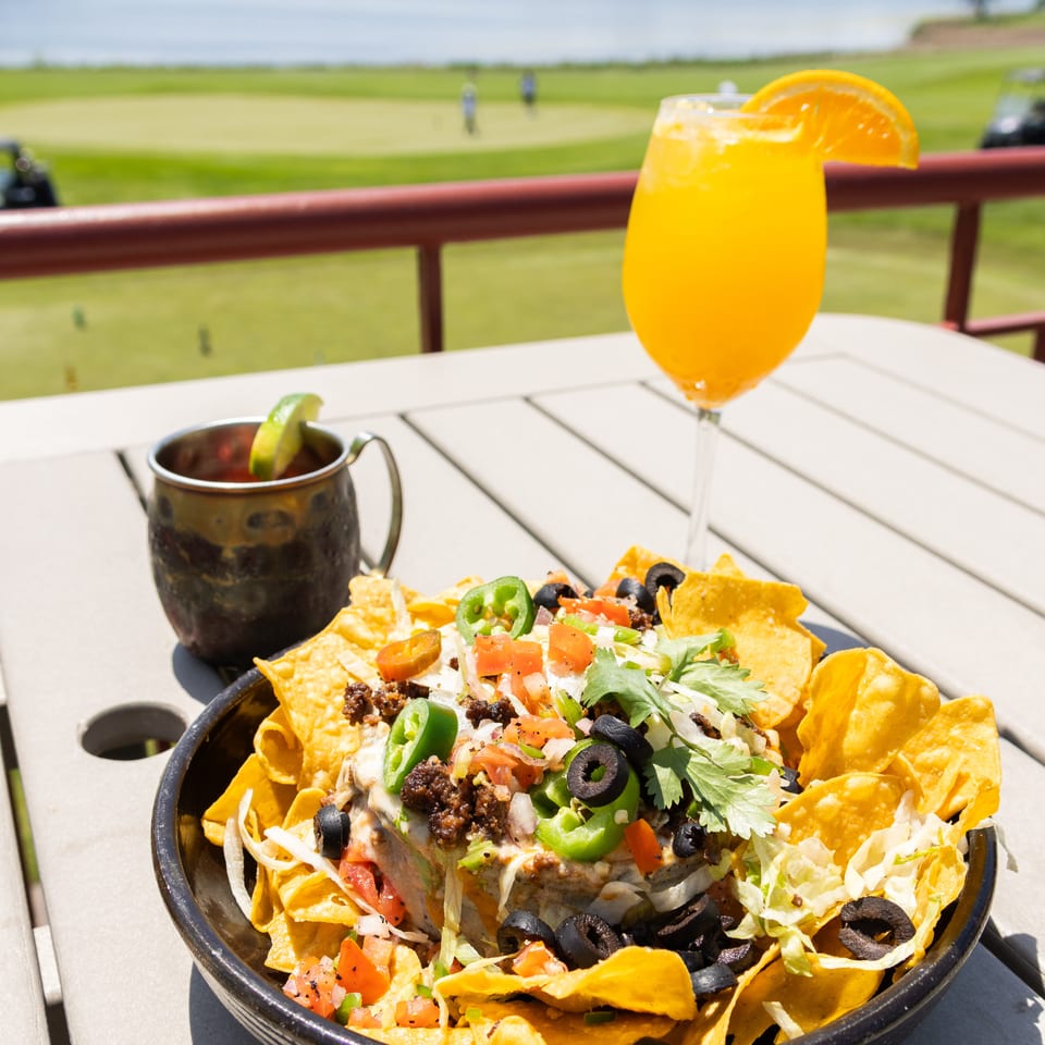 Loaded nachos with cheese, seasoned meat, jalapeños, olives, tomatoes, and lettuce beside a citrus-garnished orange drink overlooking a lakeside golf course.