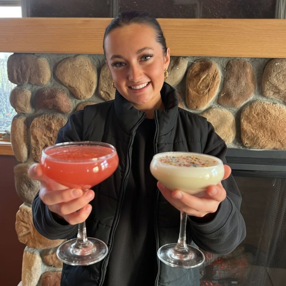 Smiling server holds red and creamy cocktails in stemmed glasses before a rustic stone fireplace.