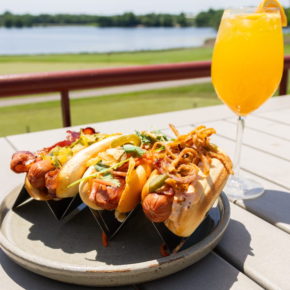 Loaded hot dogs topped with bacon, pickles, herbs, and crispy onions beside a bright orange drink overlooking a lakeside landscape.