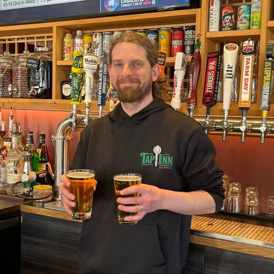 Bartender wearing a Tap Inn hoodie holds two beers in front of a bar lined with colorful beer taps.