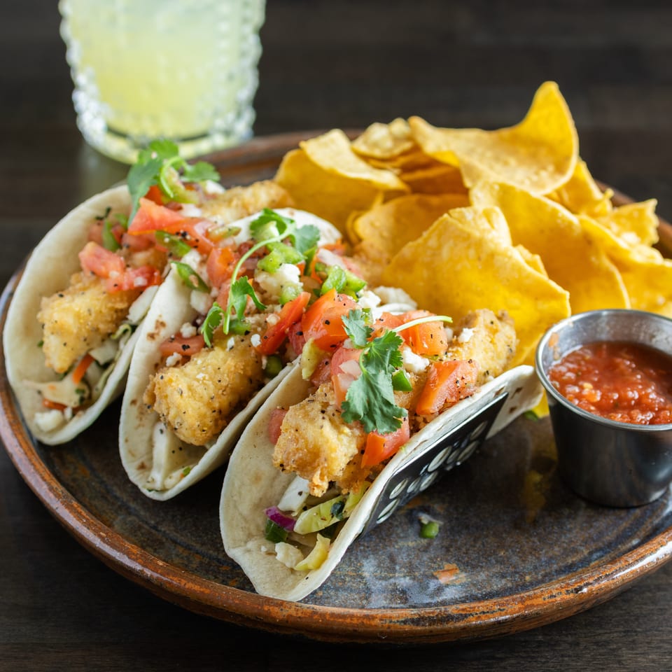 Three crispy fish tacos topped with pico de gallo and cilantro, served with tortilla chips, salsa, and a lime drink.