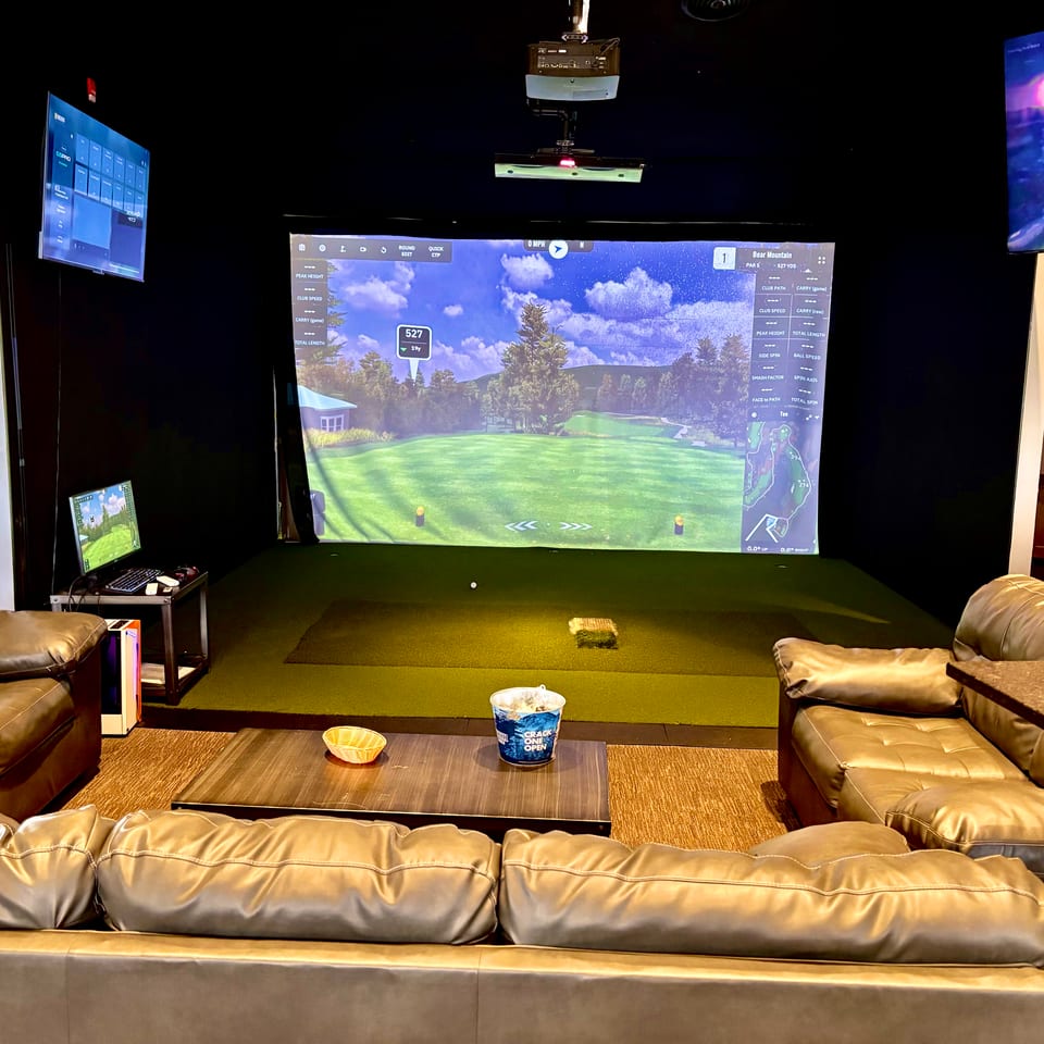 Indoor golf simulator lounge with a large projected course screen, overhead projector, leather seating, and a snack table.