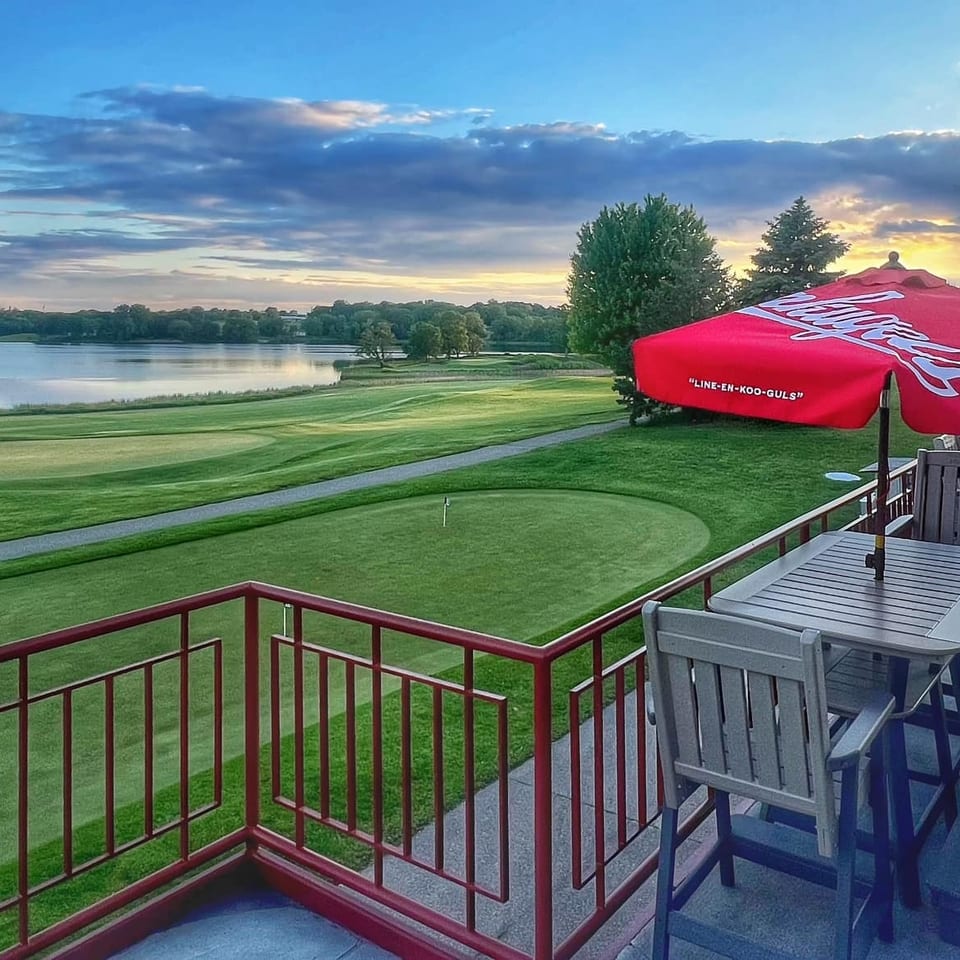Restaurant patio with red umbrellas overlooks a manicured golf course and lake beneath a colorful sunset sky.