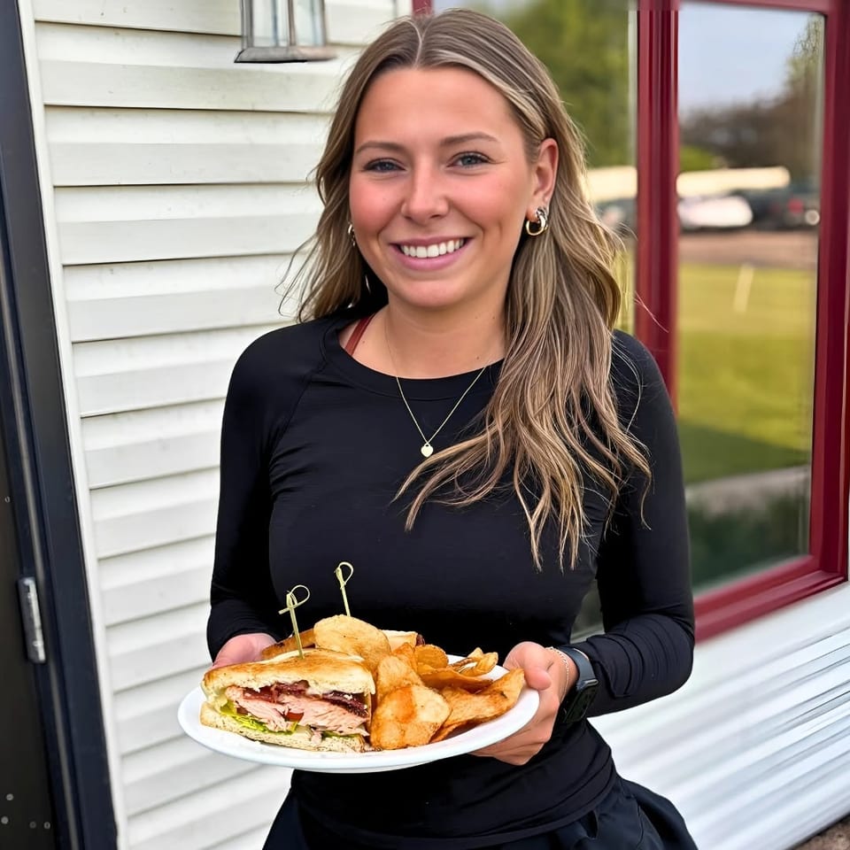 Smiling server holds a plate with a turkey sandwich, potato chips, and toasted bread outside a restaurant.