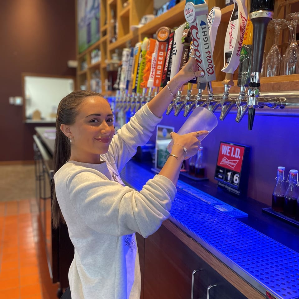 Bartender points to colorful beer taps at a warmly lit bar with wooden shelves and blue counter lighting.