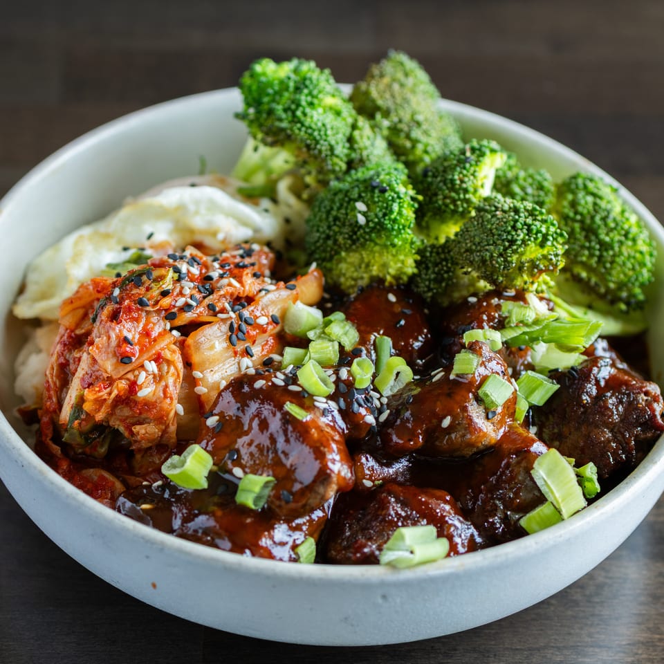 Korean-inspired bowl with glazed meat, kimchi, broccoli, fried egg, rice, sesame seeds, and scallions.