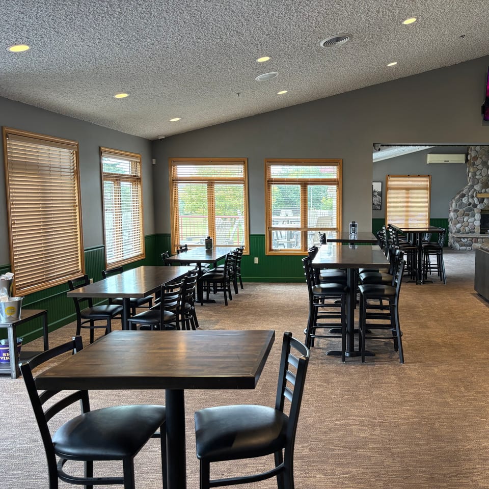 Bright restaurant dining room with wooden tables, black chairs, large windows, green wainscoting, and a stone fireplace.