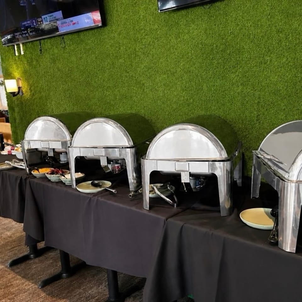 Buffet table with polished stainless-steel chafing dishes, serving utensils, plates, and bowls against a green textured wall.