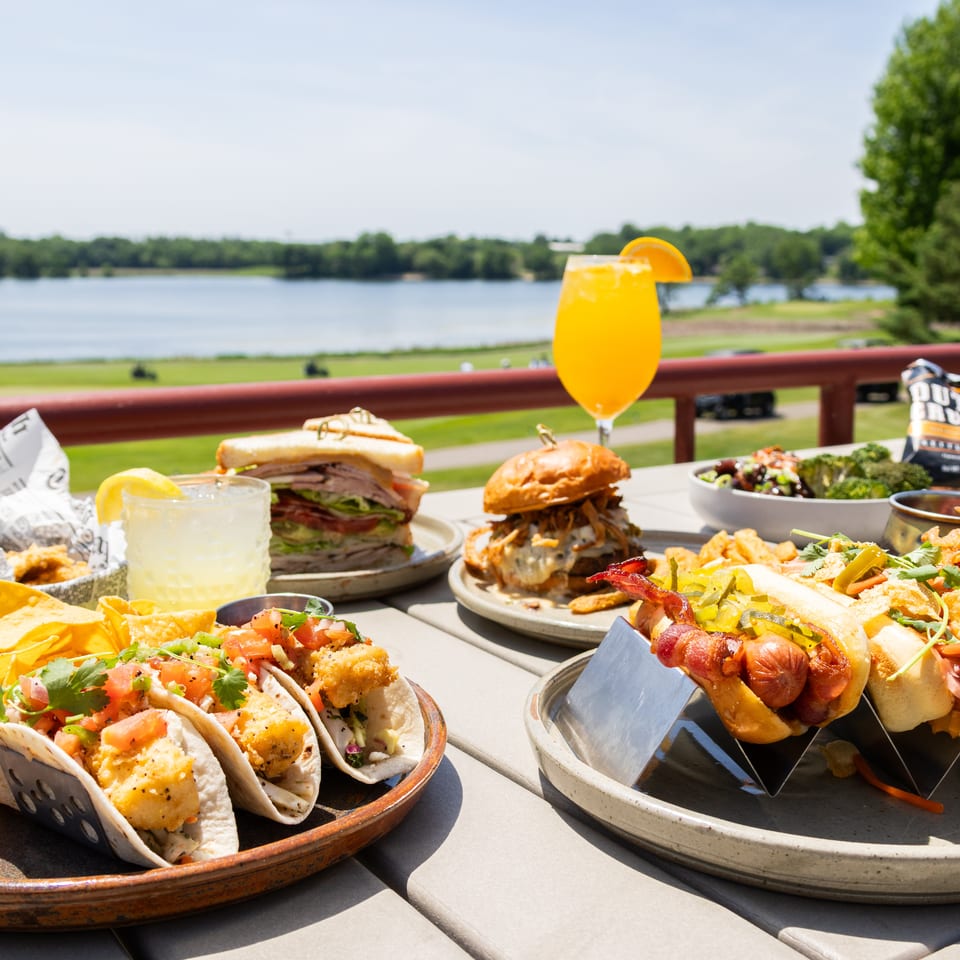 Outdoor restaurant table overlooking a lake, filled with tacos, sandwiches, burgers, loaded hot dogs, sides, and orange drinks.