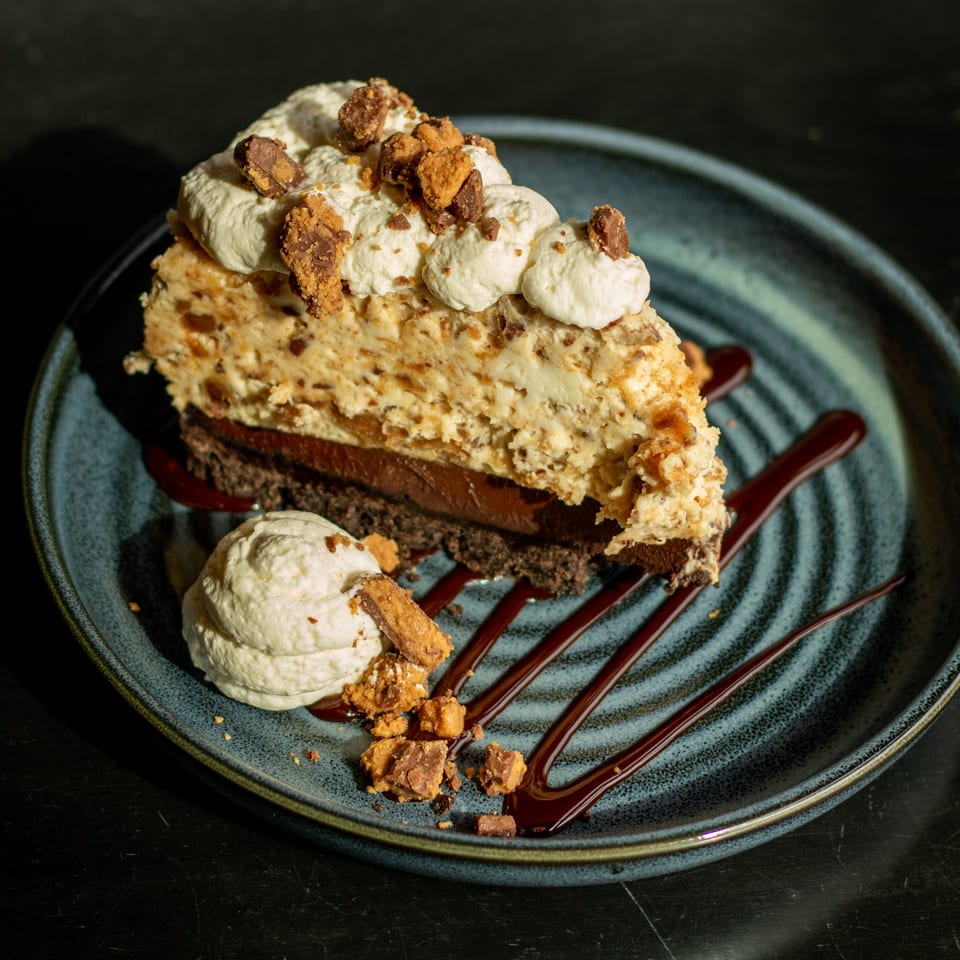 Slice of layered chocolate and nut dessert topped with whipped cream, crushed candy pieces, and chocolate sauce on a blue plate.