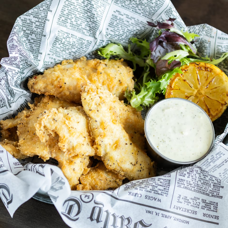 Basket of golden fried fish with mixed greens, grilled lemon, and creamy dipping sauce on newspaper-lined paper.