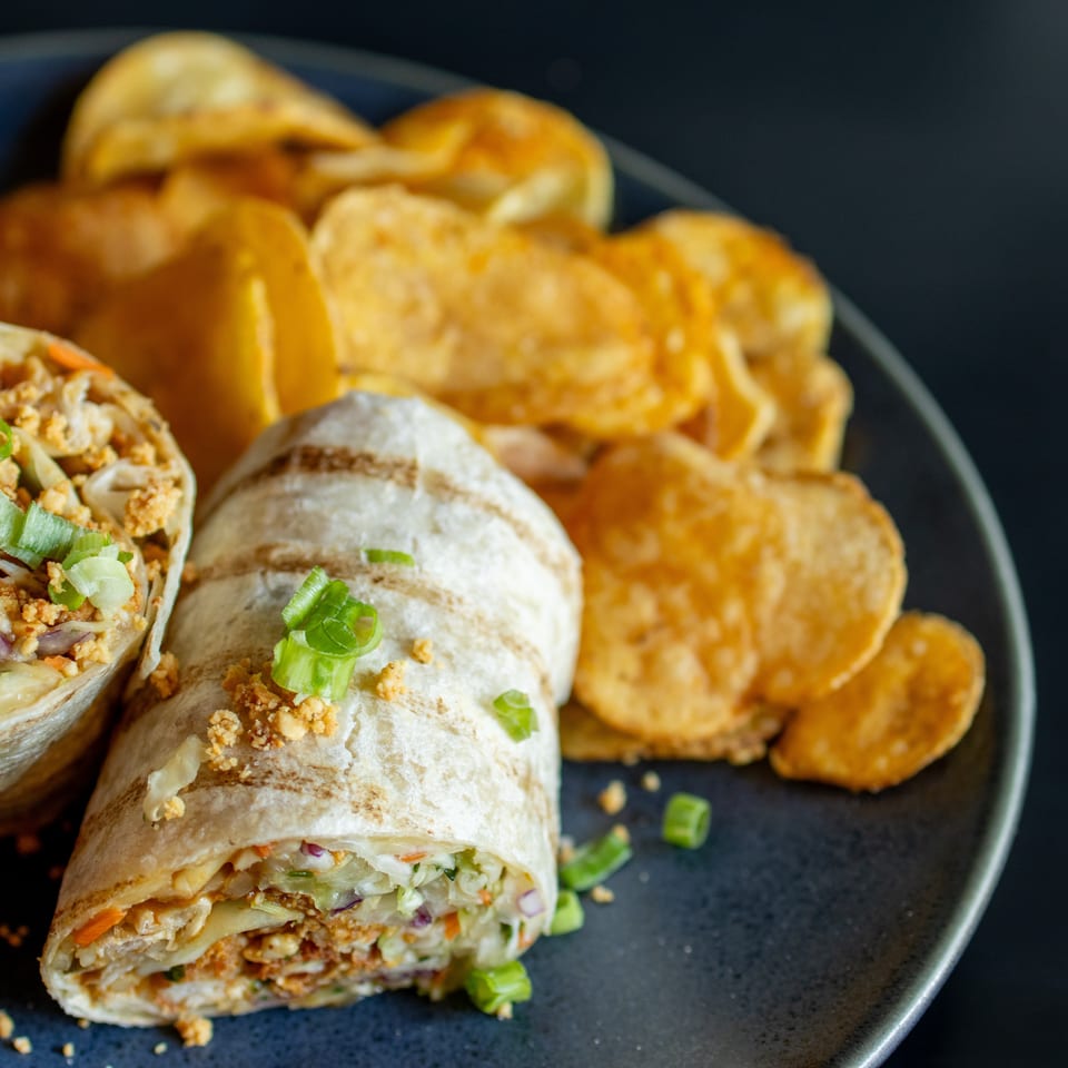 Sliced vegetable-filled wraps with savory crumbles, creamy dip, and golden tortilla chips arranged on a dark plate.