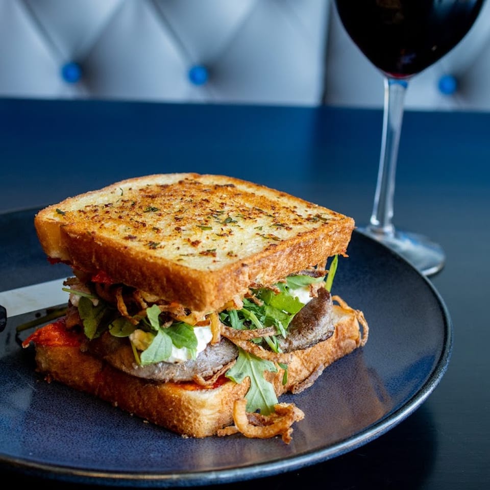 Gourmet sandwich with steak, arugula, and crispy onions on toasted bread, served with red wine.