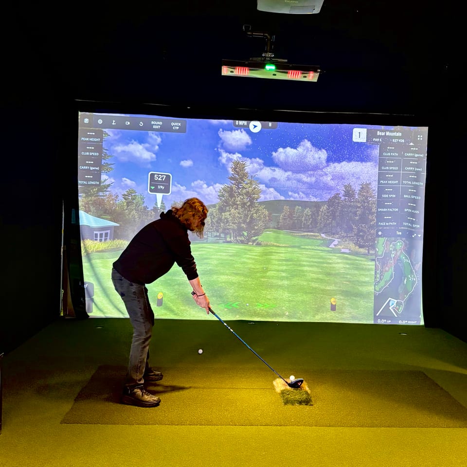 Person prepares to hit a golf ball in an indoor simulator bay displaying a virtual mountain course.