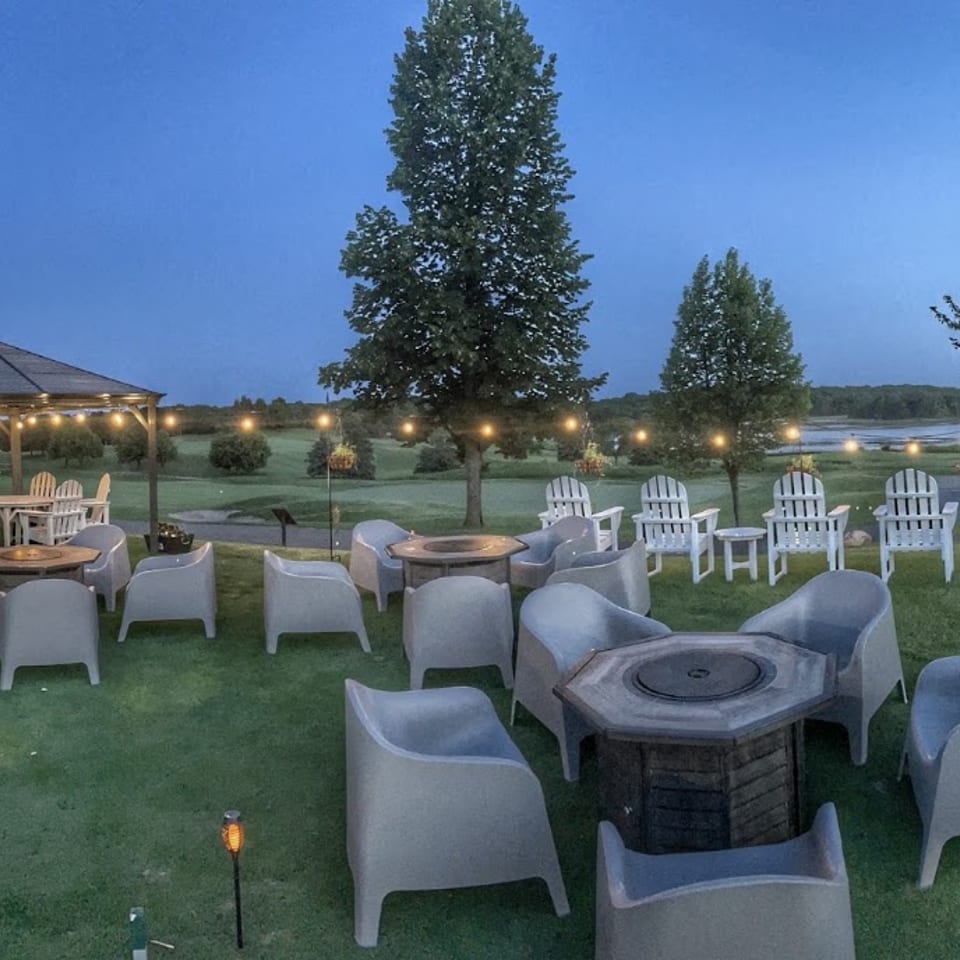 Outdoor patio at dusk with a gazebo, string lights, fire pits, and seating overlooking a lake.