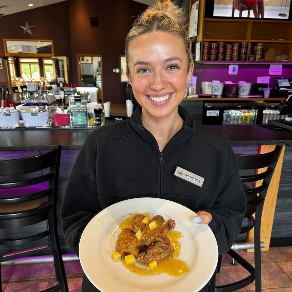 Smiling restaurant server holds a plate of glazed chicken pieces topped with diced fruit inside a warmly lit bar area.
