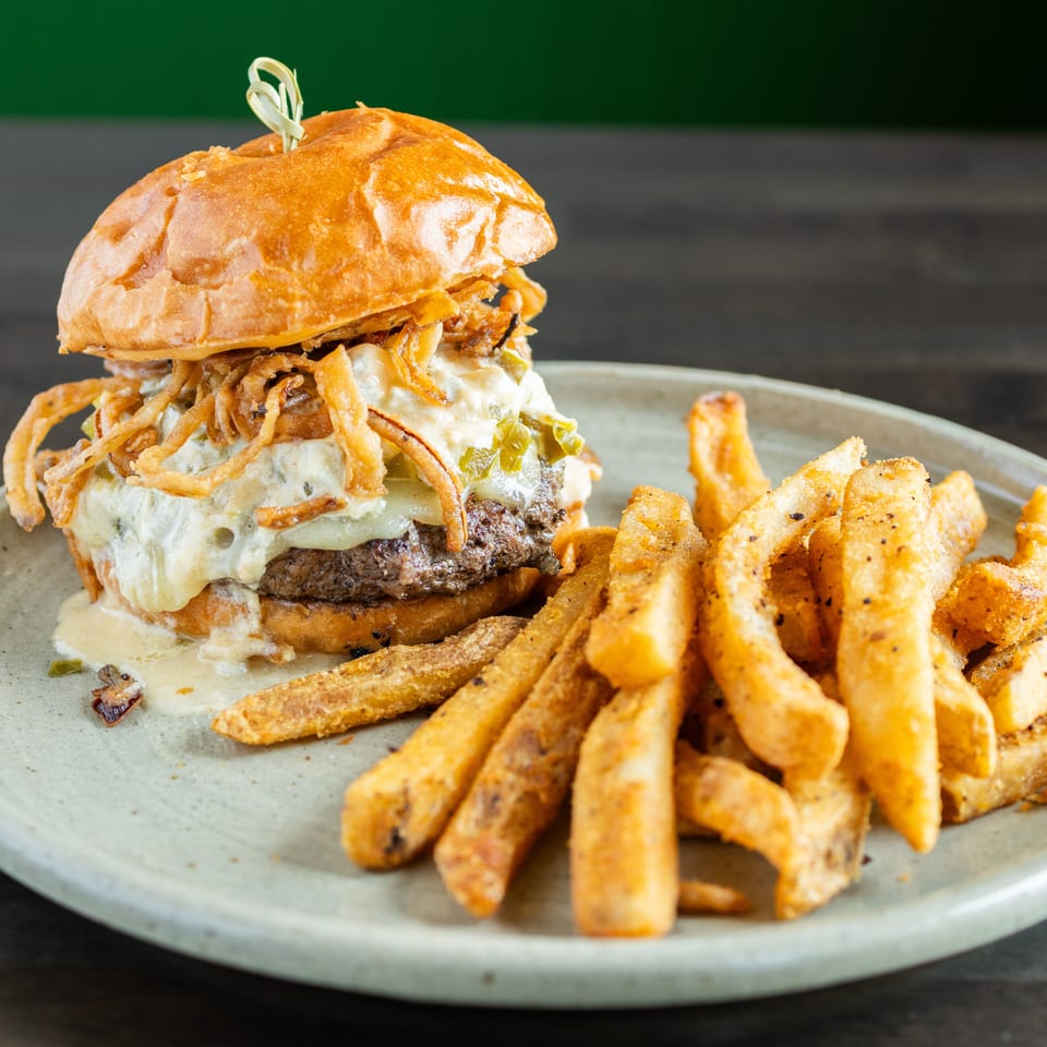 Cheeseburger topped with crispy fried onions and sauce, served with golden seasoned fries on a ceramic plate.