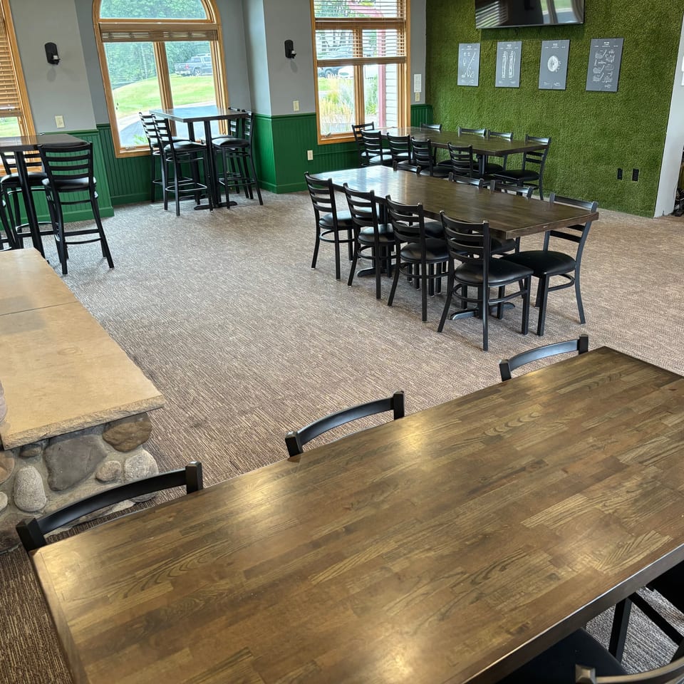 Spacious restaurant dining area with wooden tables, black chairs, green walls, large windows, and a mounted television.