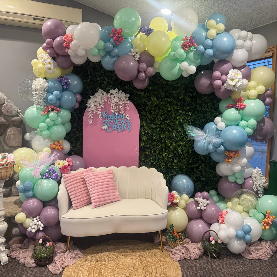 Pastel balloon and floral Easter display surrounding a white loveseat, with a pink Happy Easter sign and decorative baskets.