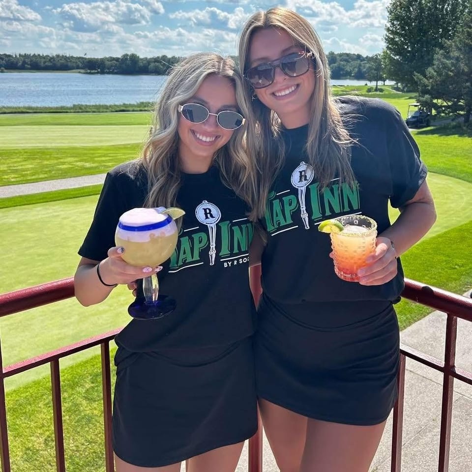 Two smiling women in Tap Inn shirts hold colorful cocktails on a balcony overlooking a golf course and lake.